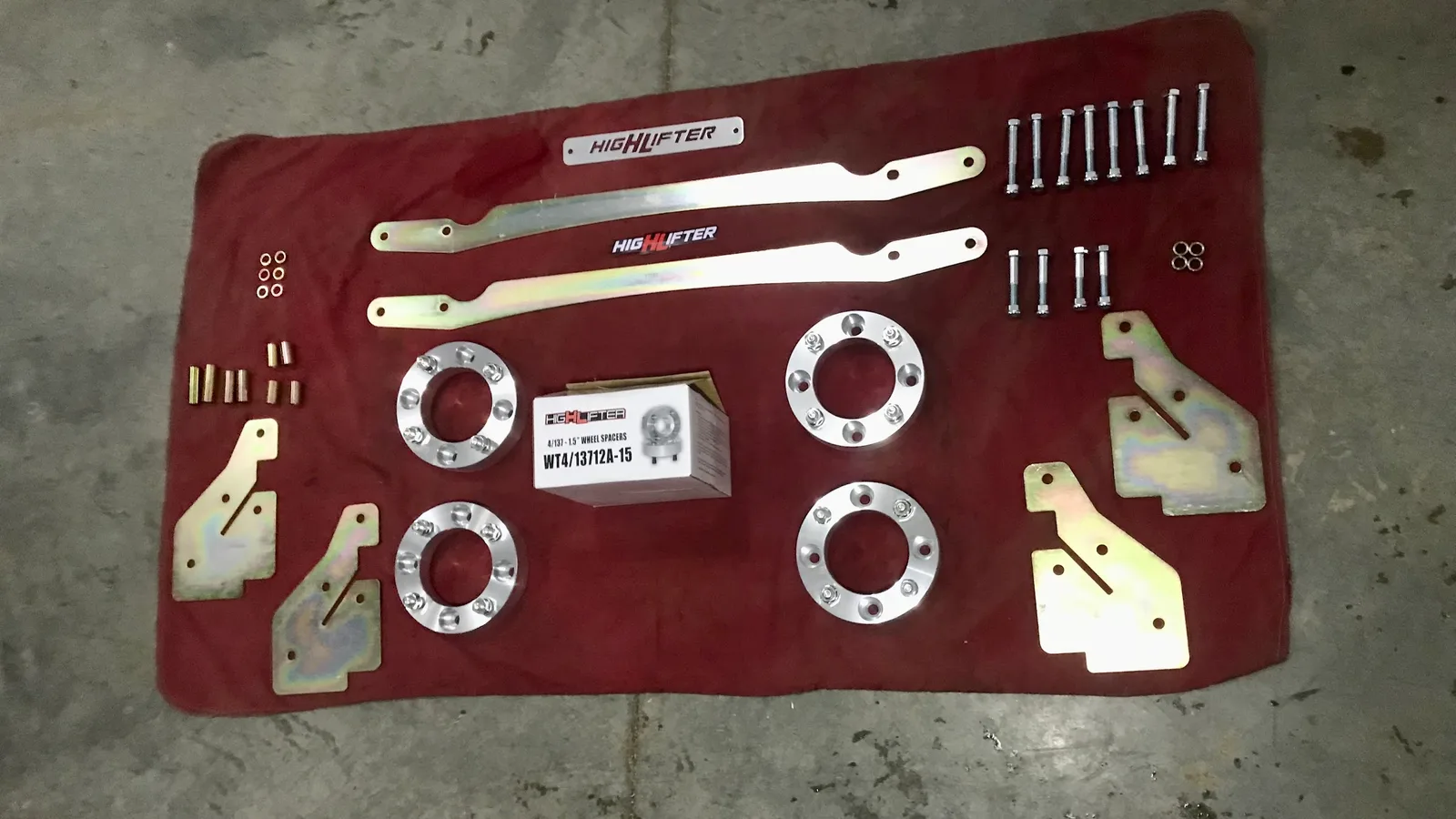 A High Lifter ATV lift kit laid out in rows on a red shop blanket on the concrete shop floor: brackets, four wheel spacers, sorted bolts and the parts box